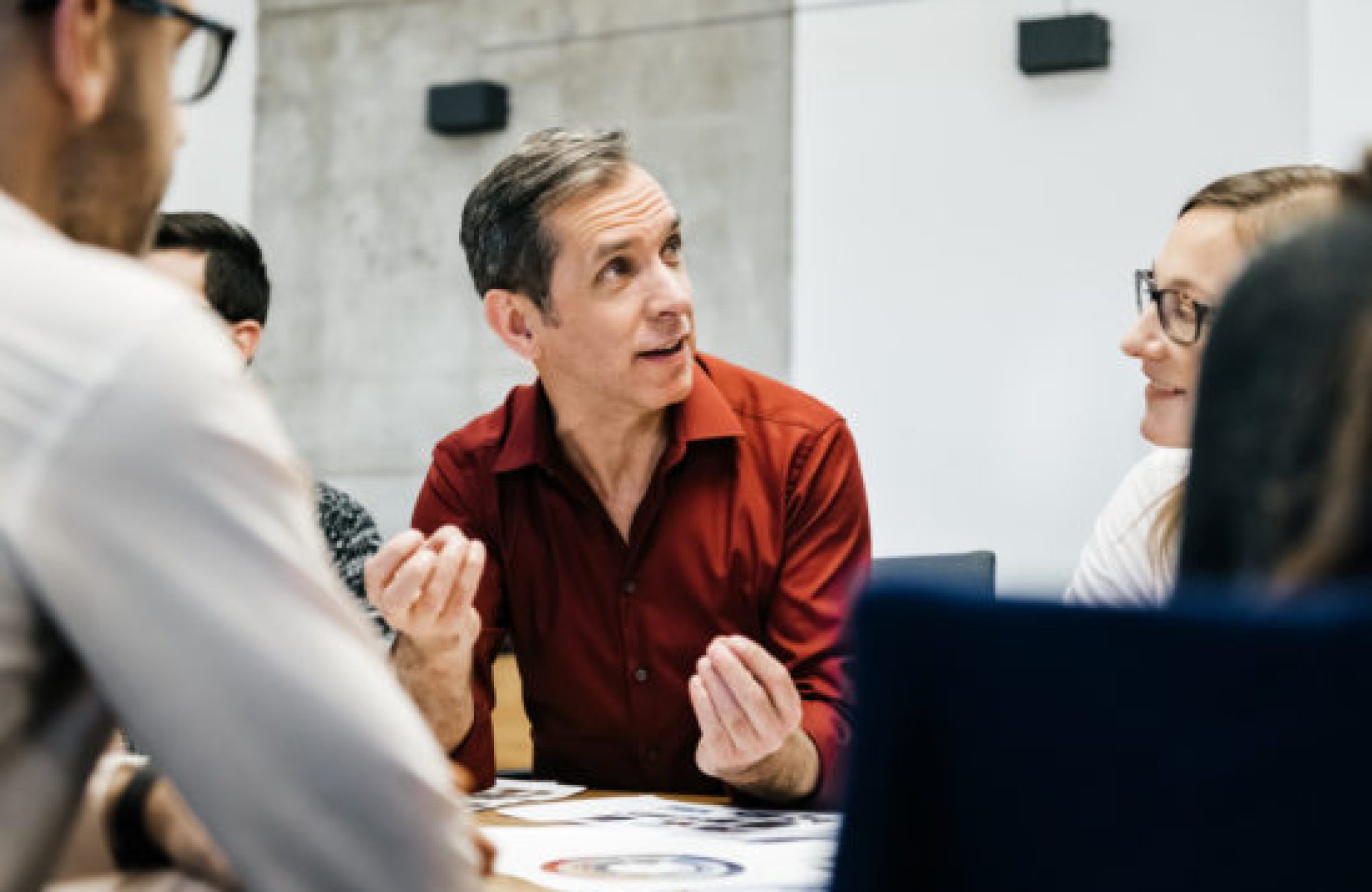 A caucasian man is speaking in a business meeting surrounded by colleagues listening to him. He uses his hands and seems convinced and focused.